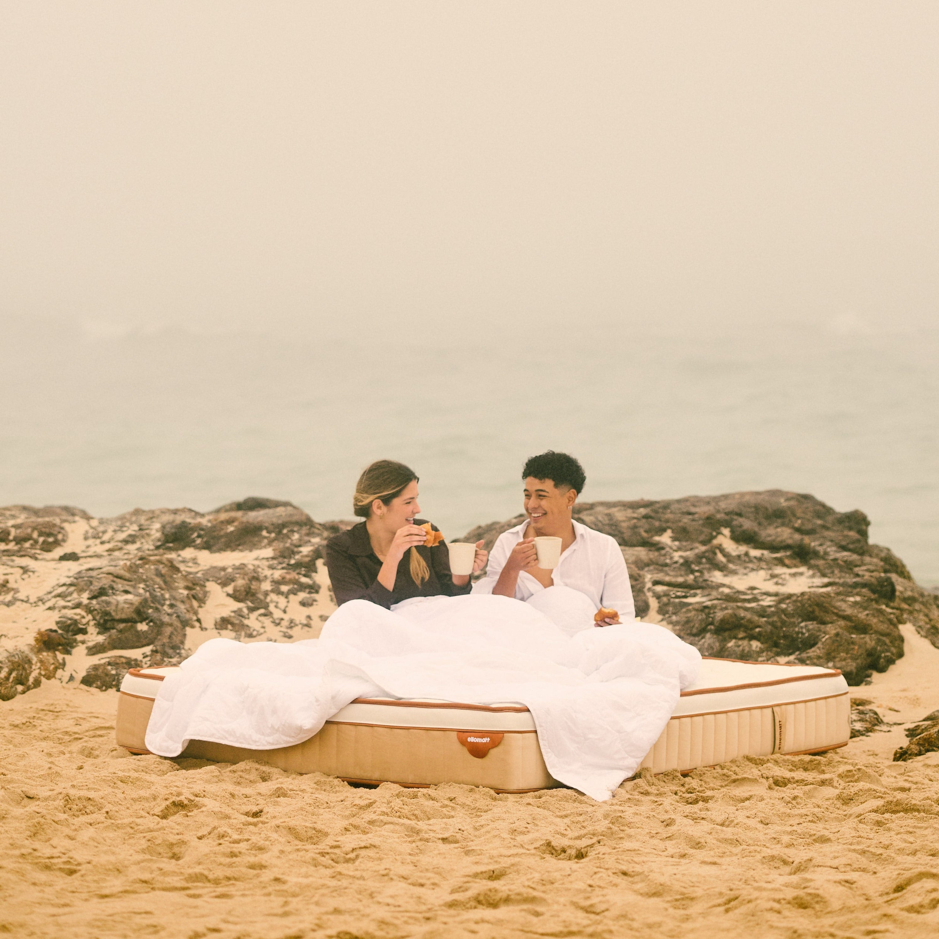 Two people sitting on a mattress in the sand with a scenic background